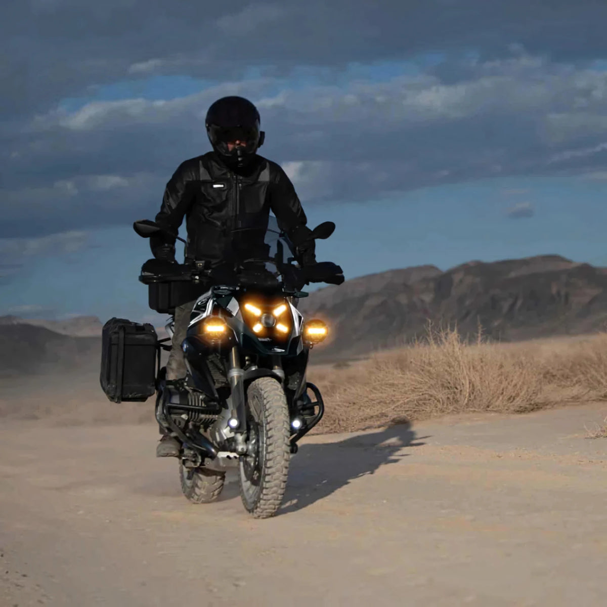 Rider on a BMW R 1300 GS motorcycle with bright auxiliary LED driving lights on a dusty desert trail.