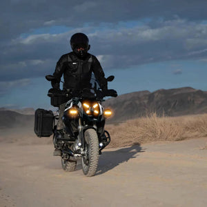 Rider on a BMW R 1300 GS motorcycle with bright auxiliary LED driving lights on a dusty desert trail.